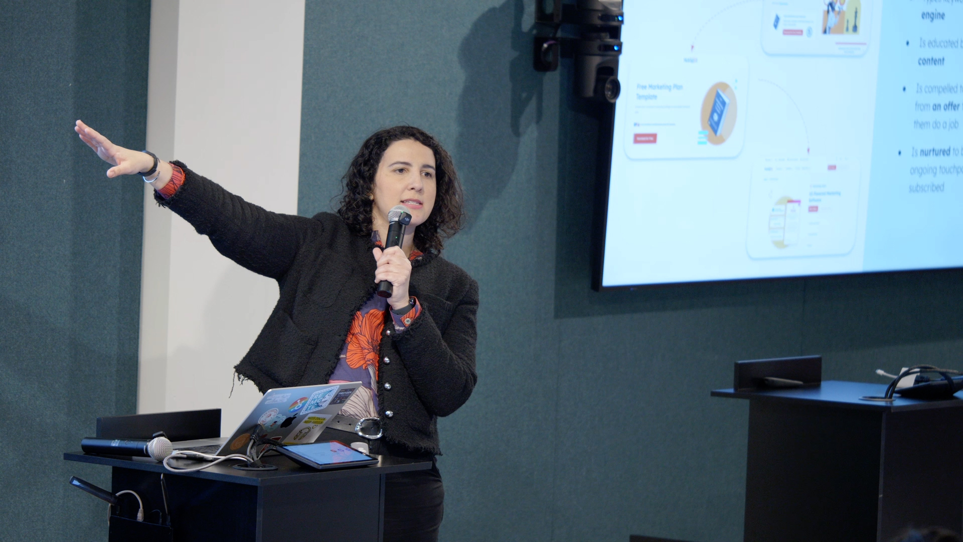 A woman with curly hair speaks into a microphone while gesturing toward a marketing presentation screen at a conference.