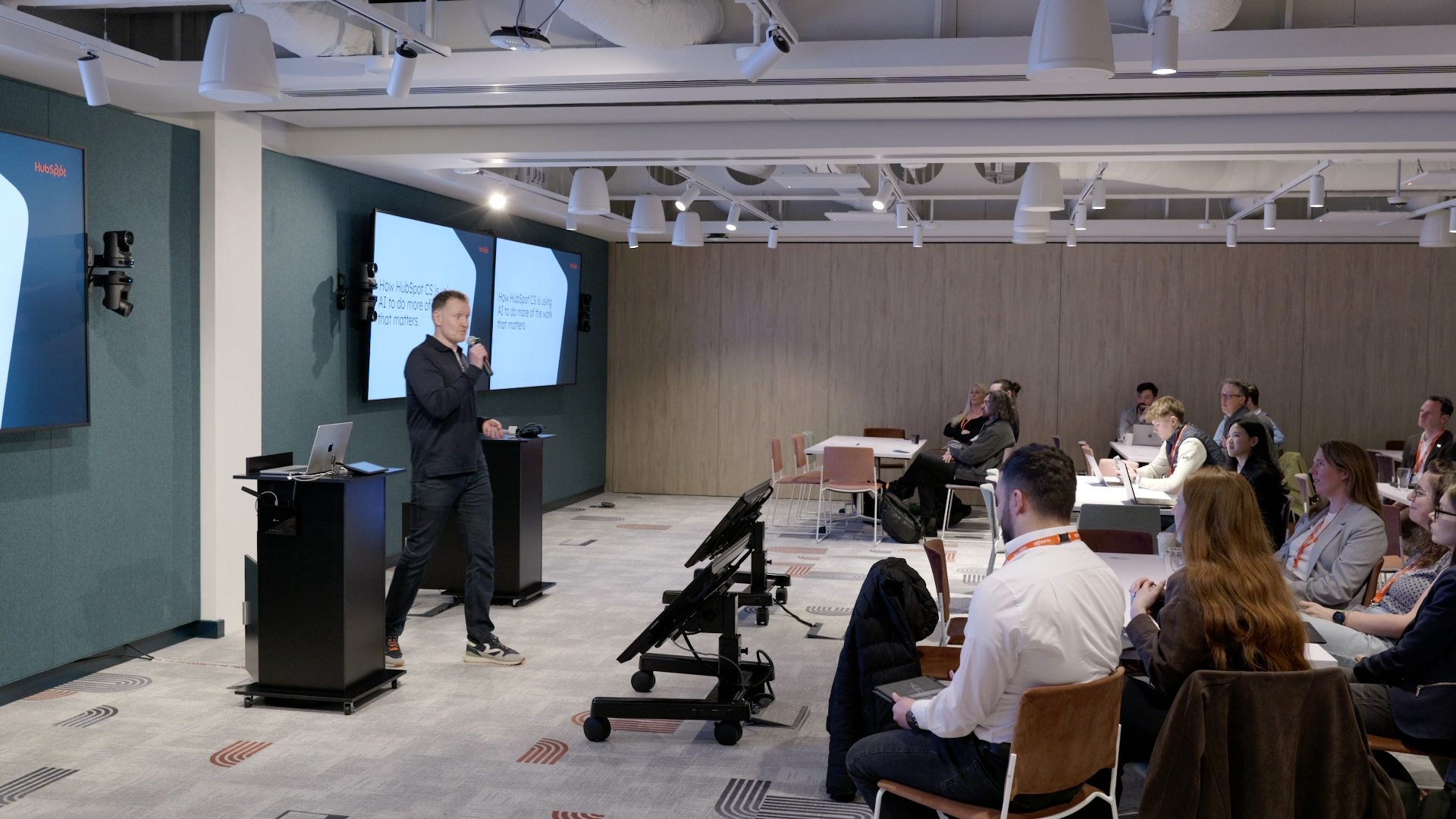 A presenter speaks at a HubSpot conference about AI in customer success, addressing a seated audience.