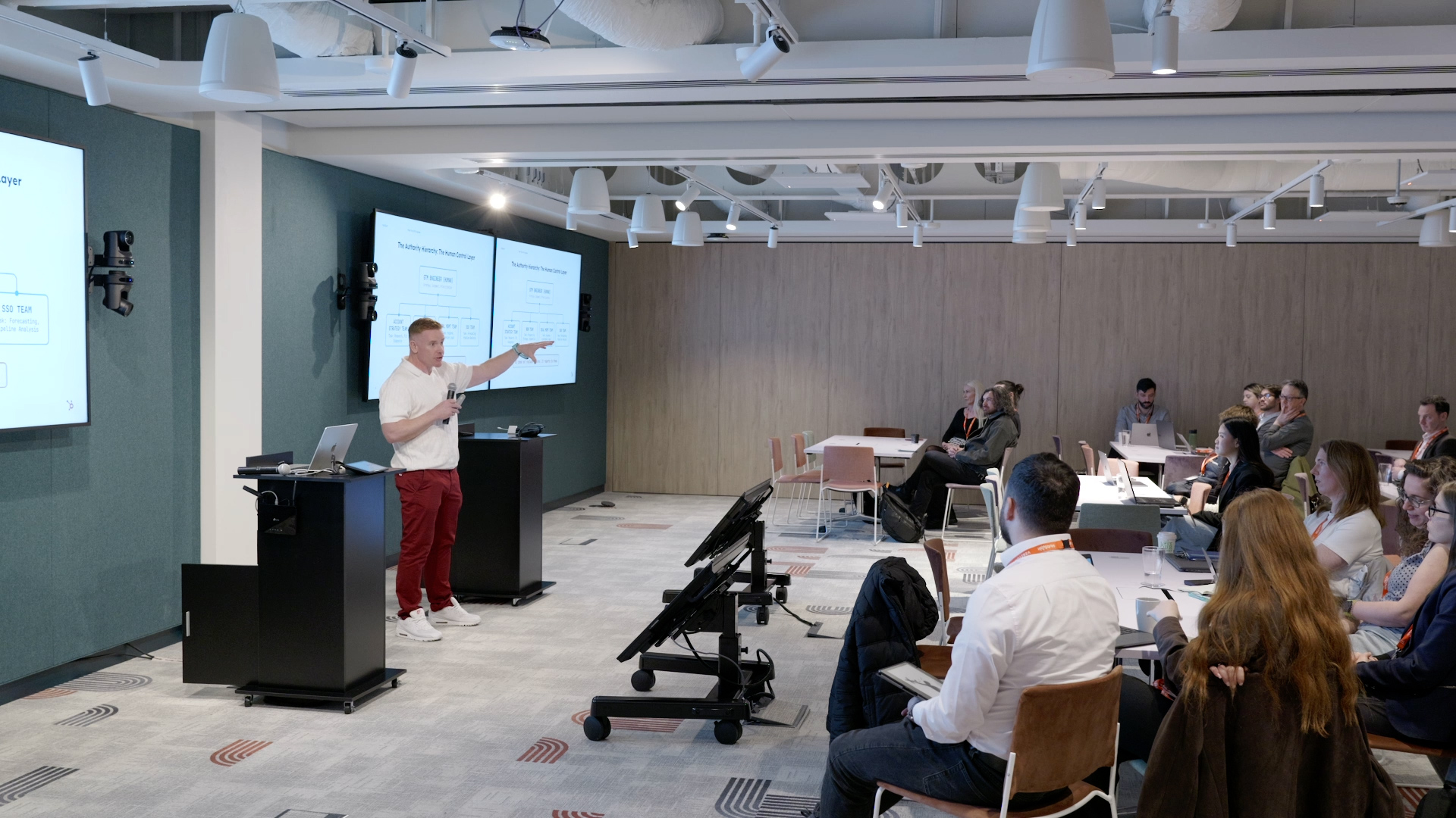 A presenter in a white shirt gestures toward dual screens while addressing an audience in a modern conference room.