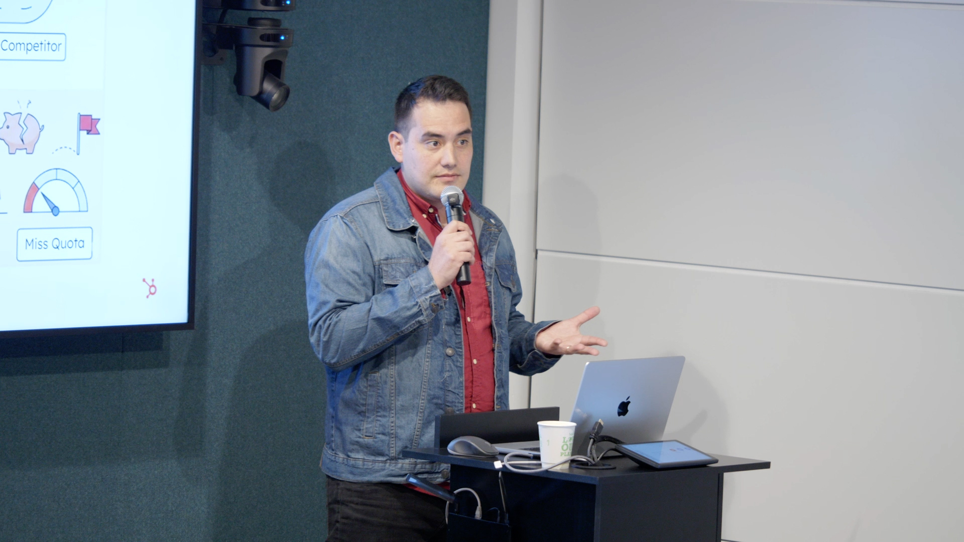 A man in a denim jacket presents with a microphone beside a laptop, with business slides showing competitor and quota metrics.