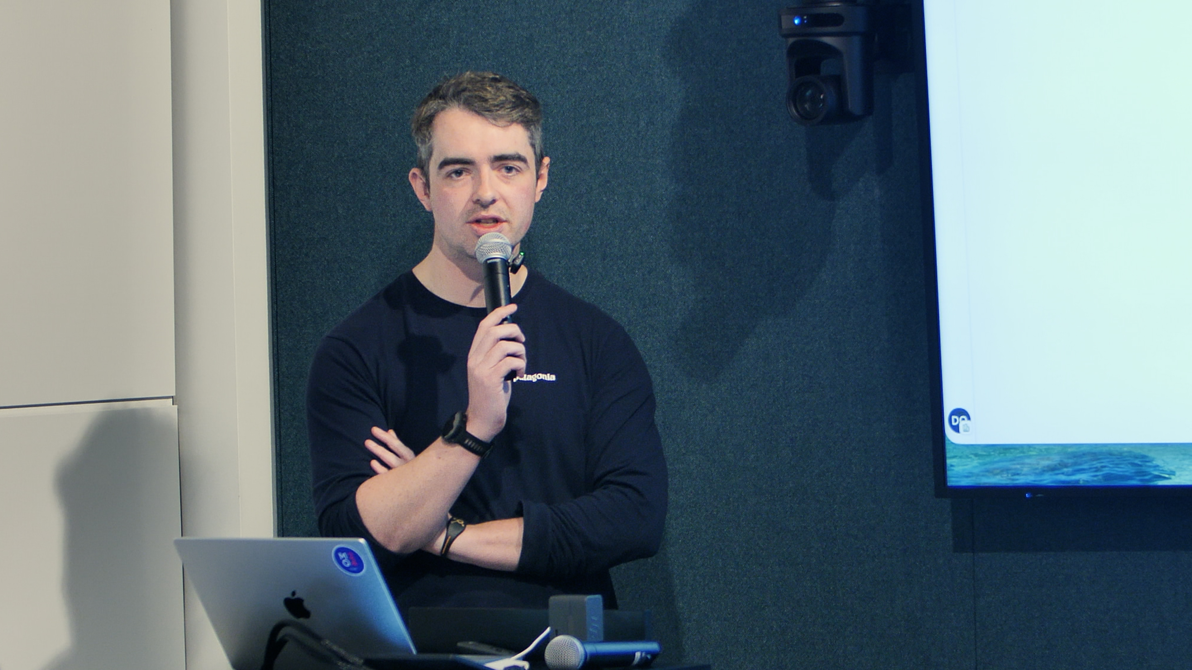 A young man in a black Patagonia shirt speaks into a microphone during a presentation with a laptop nearby.