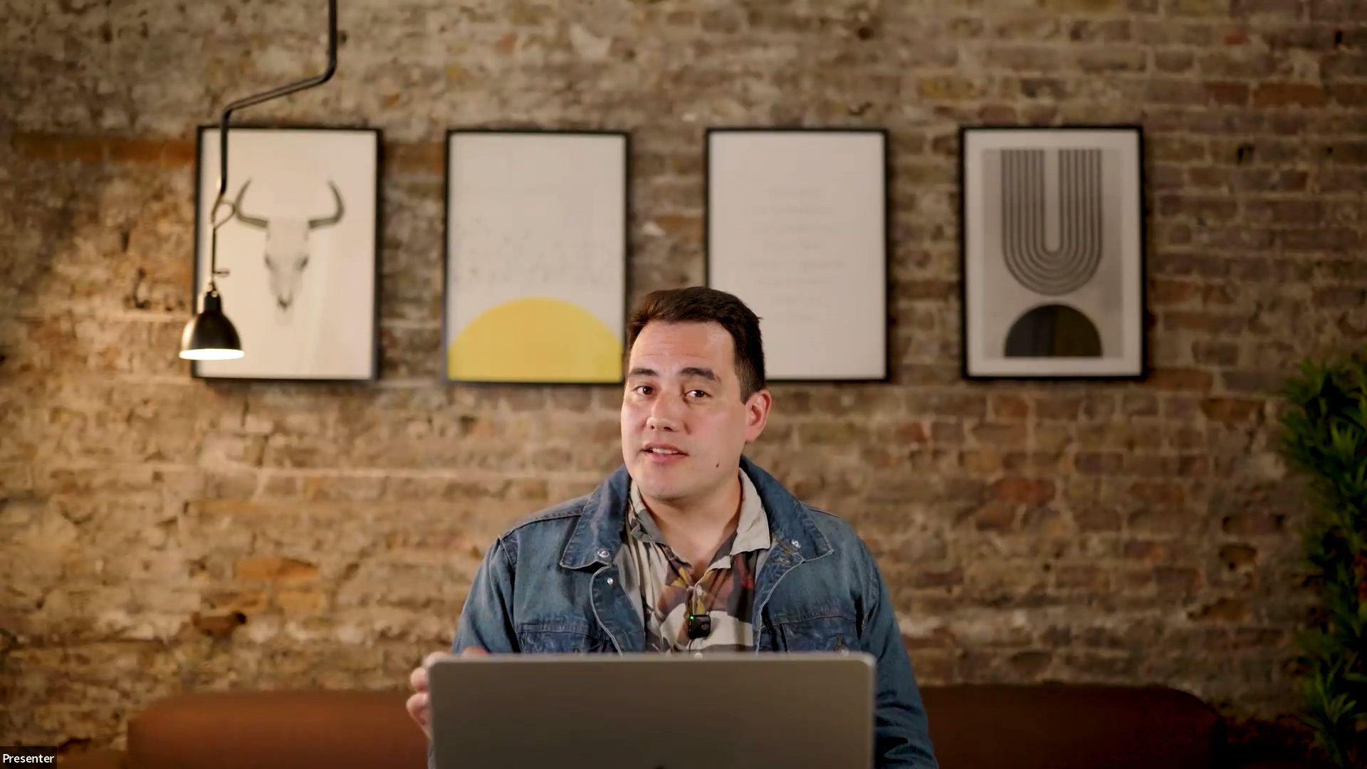 A man in a denim jacket presents from a laptop in a modern brick-walled office space.