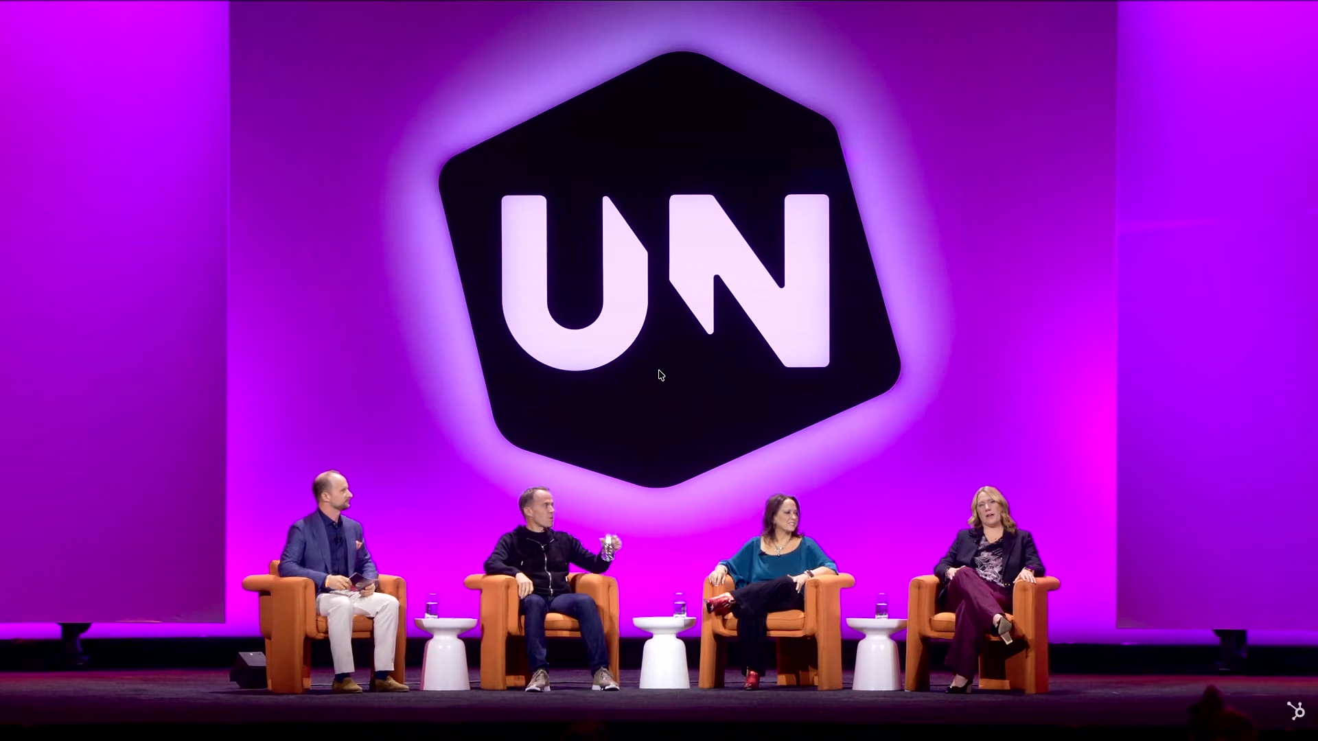 Stage panel with four people seated in orange armchairs against a pink and purple backdrop