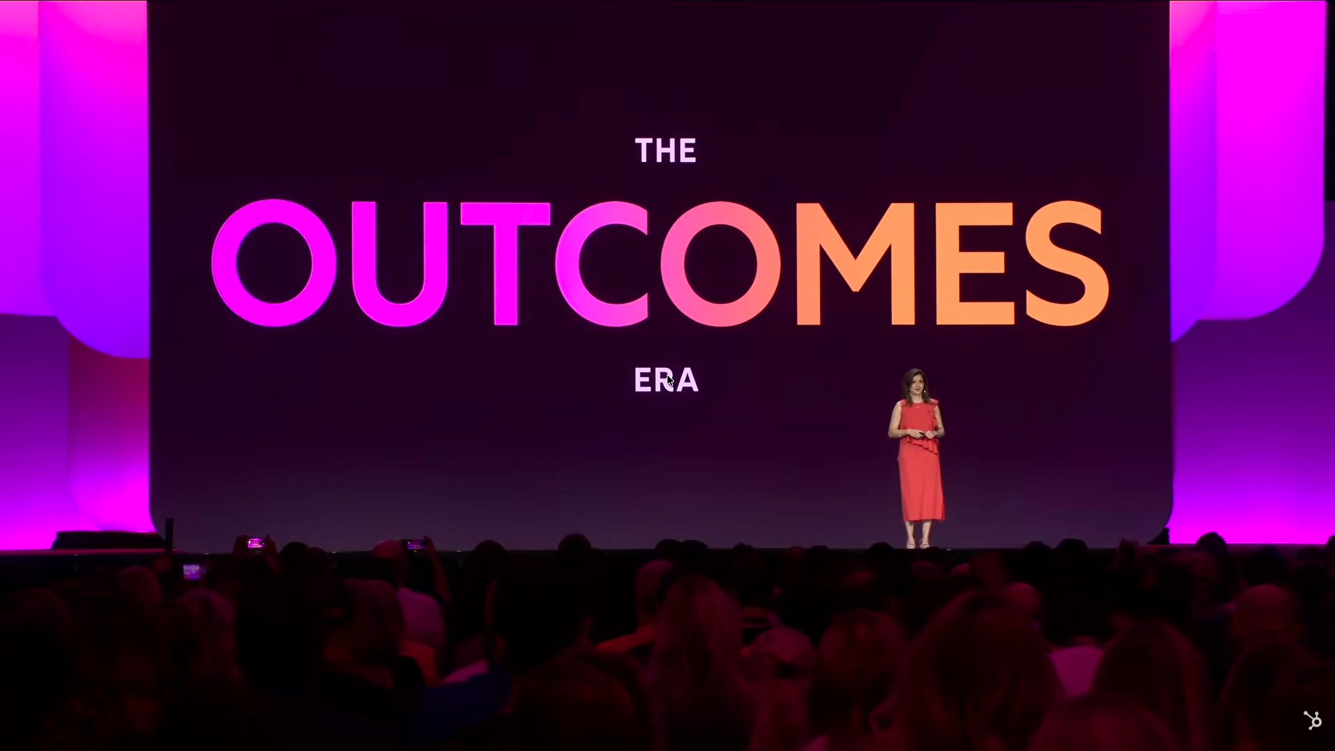 Keynote stage with a large screen displaying the words THE OUTCOMES ERA in bold colourful letters