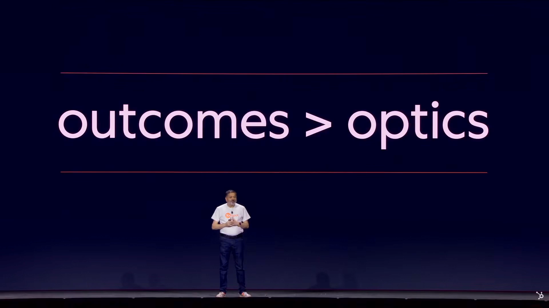 Keynote slide reading ‘outcomes > optics’ in large text, with the presenter standing on stage in front of it.