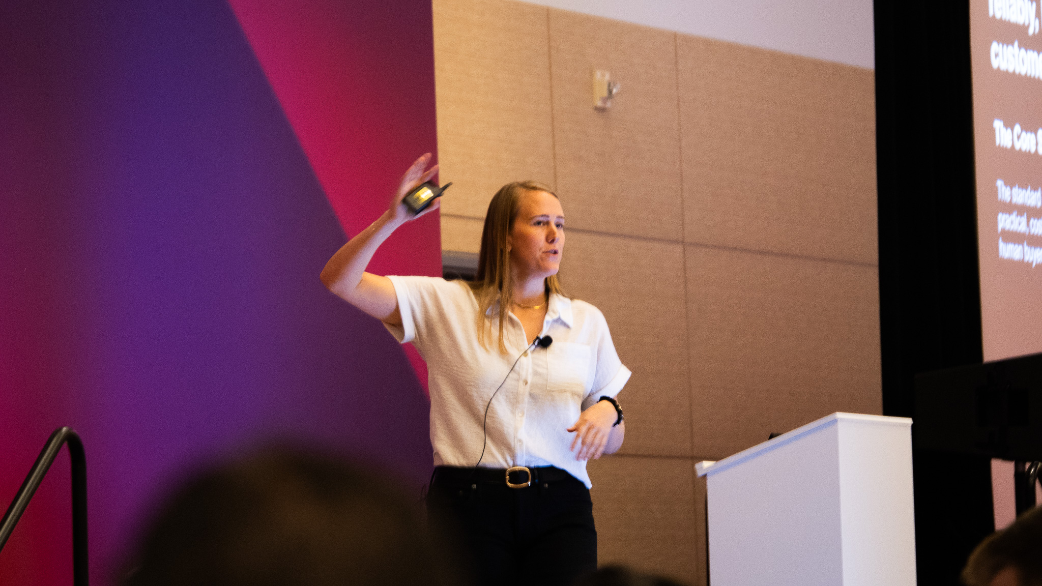 A woman stands on a stage giving a presentation, wearing a white short-sleeve shirt and dark pants with a microphone clipped to her shirt.