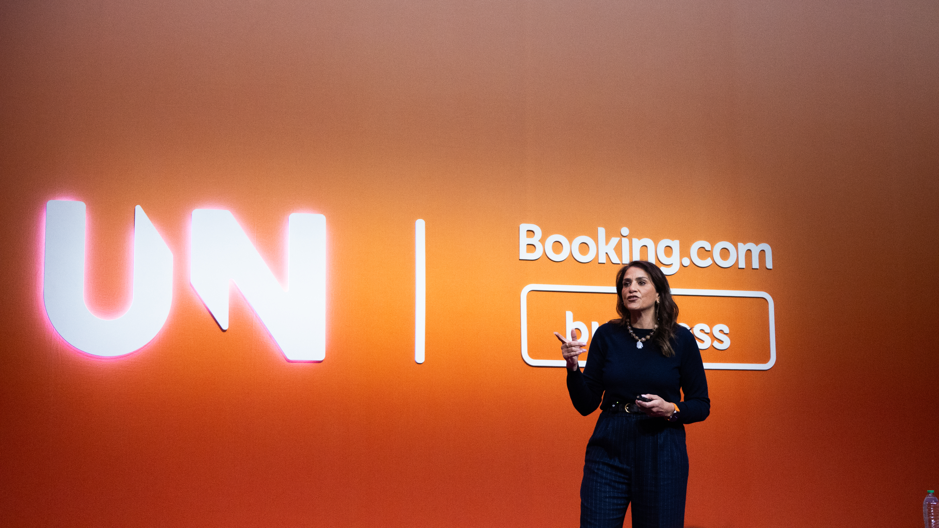 A person stands on stage in front of a large orange backdrop displaying the text 'UN' on the left and 'Booking.com business' on the right.