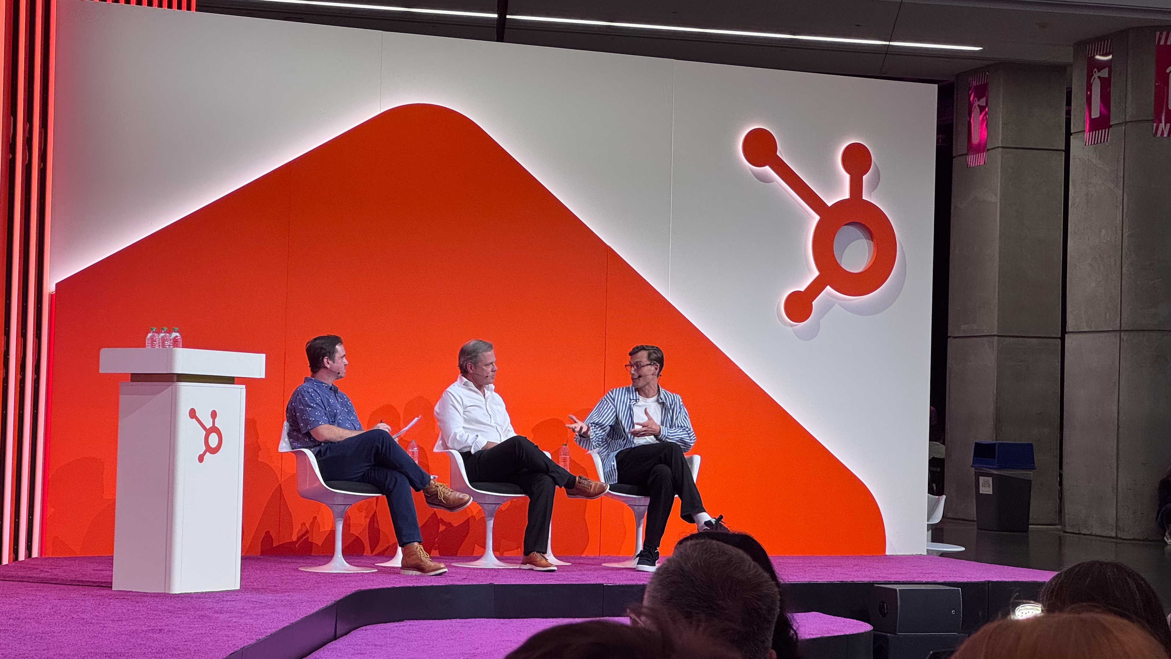 Panel discussion on stage with a bright orange and white backdrop featuring the HubSpot logo, with three people seated.