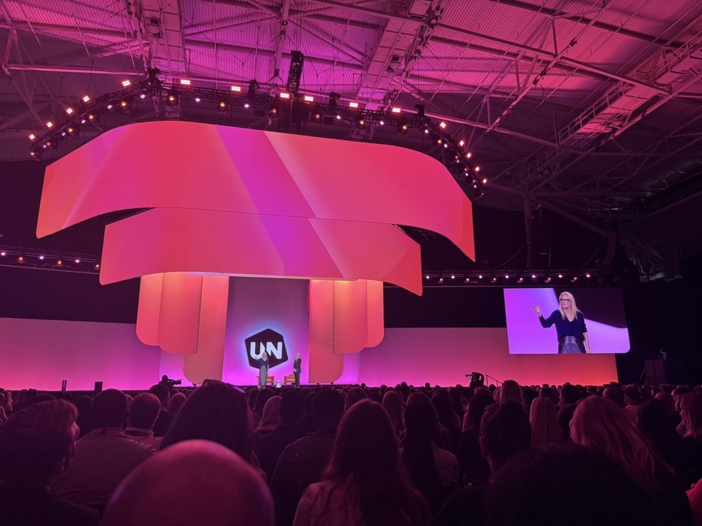 A large indoor event with a stage lit in pink and purple tones and a modern curved multi-layered screen backdrop above the audience