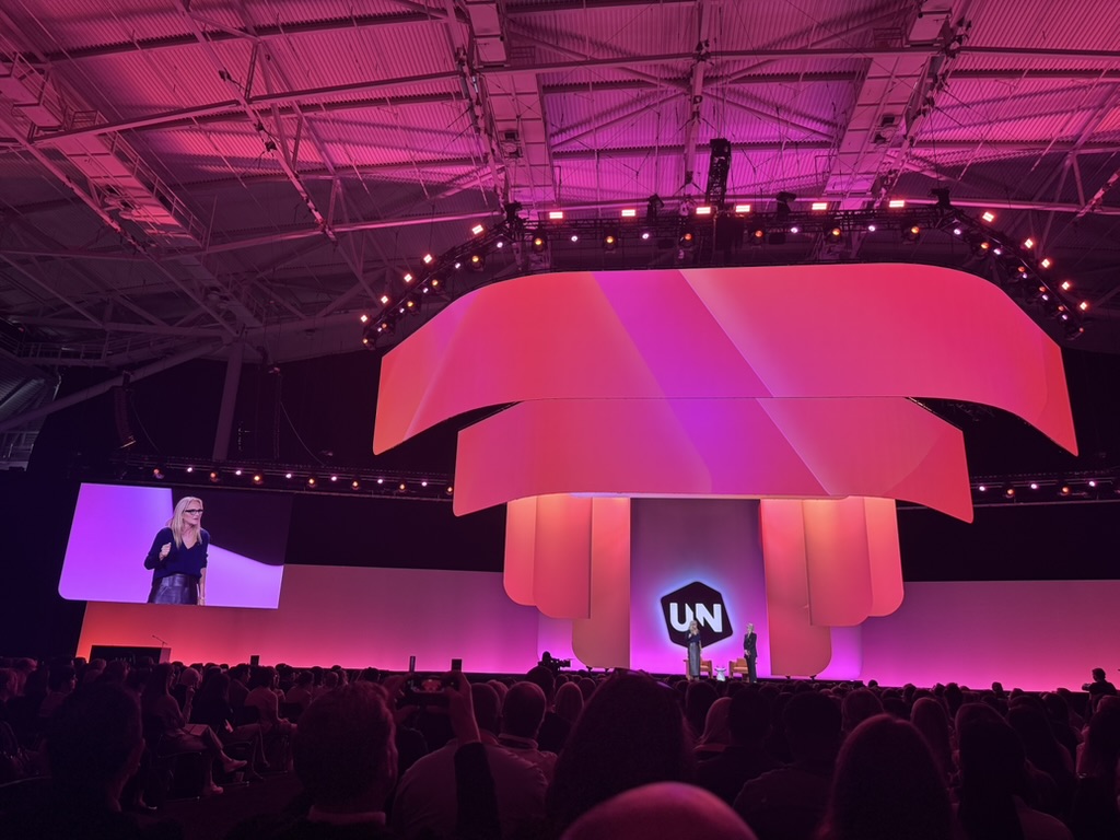 A large indoor event or conference with a stage lit in pink and purple hues, featuring a modern curved multi-tiered backdrop