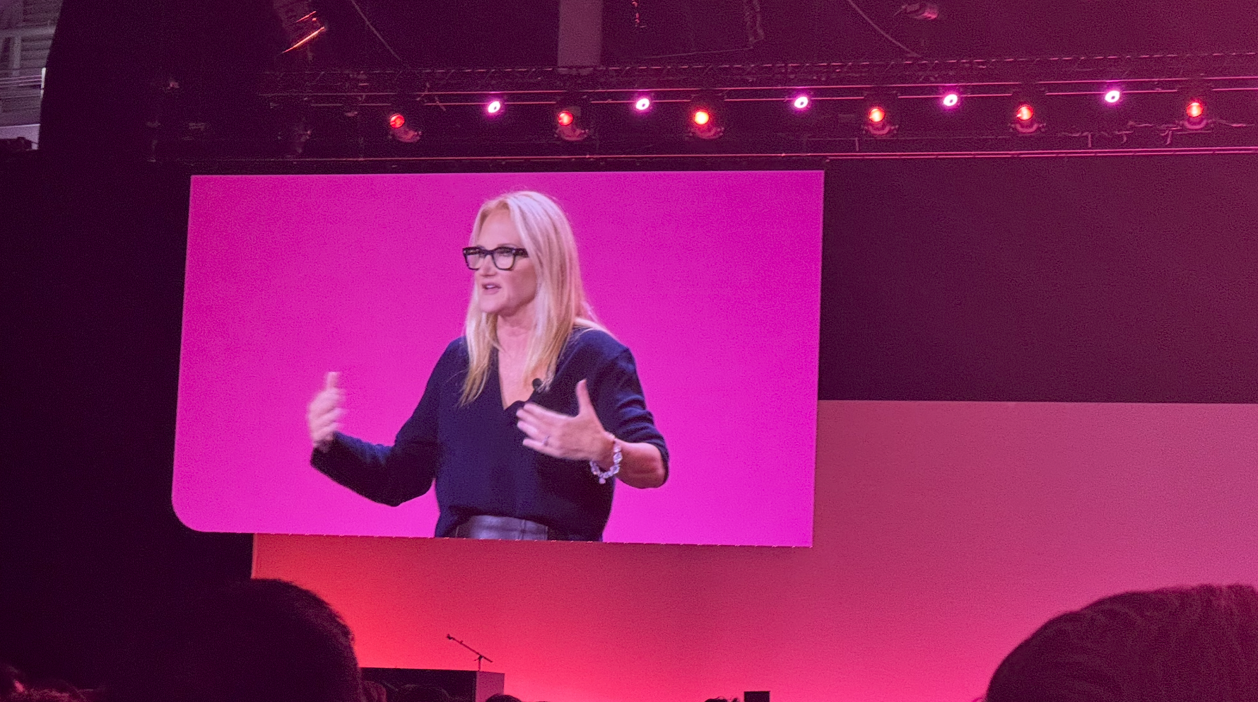 A large stage presentation with a big screen displaying the speaker, a woman with long blonde hair, mid-talk