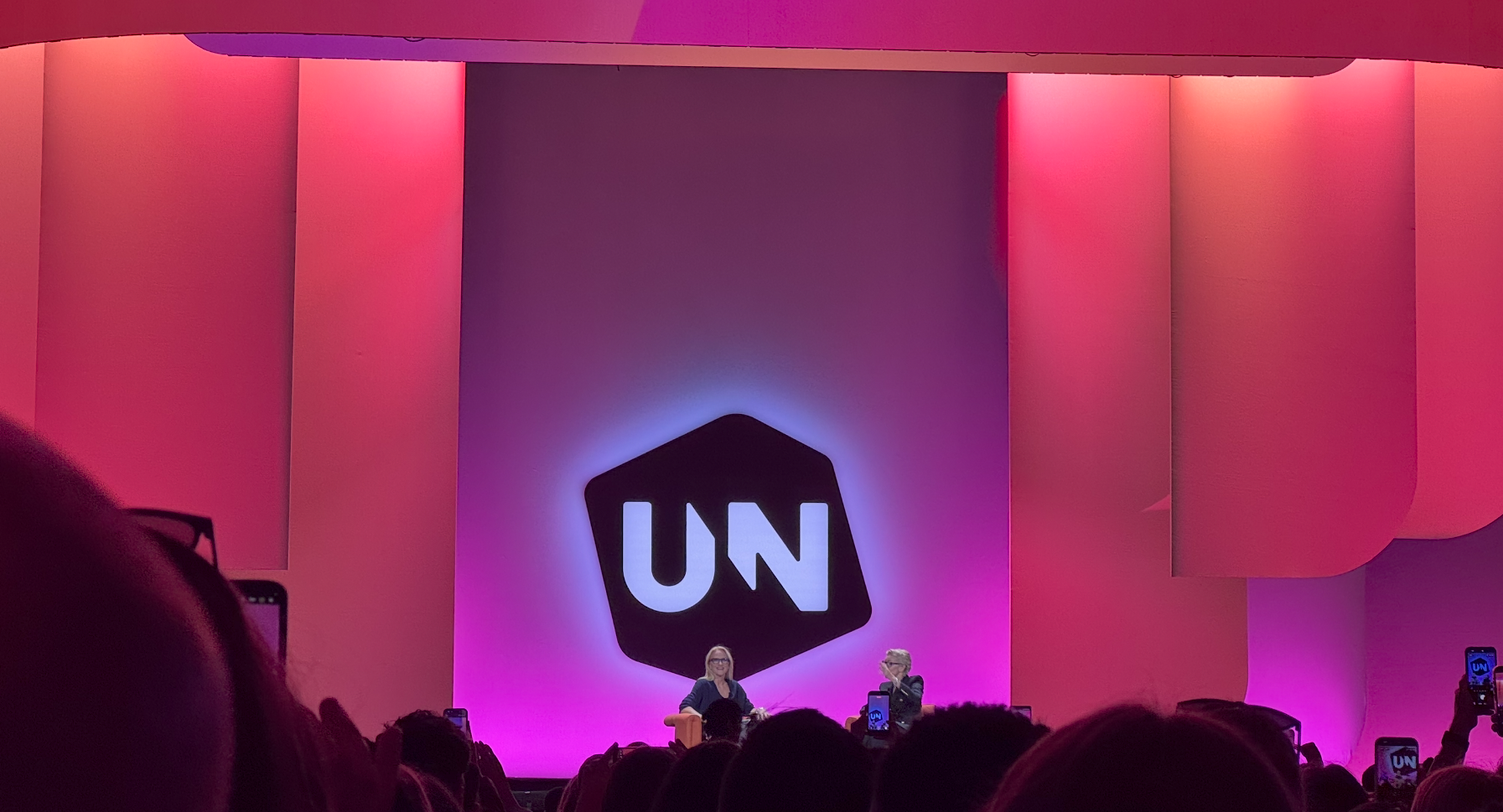 A stage with a large illuminated logo reading UN on a pink and purple lit backdrop, with two people seated in conversation