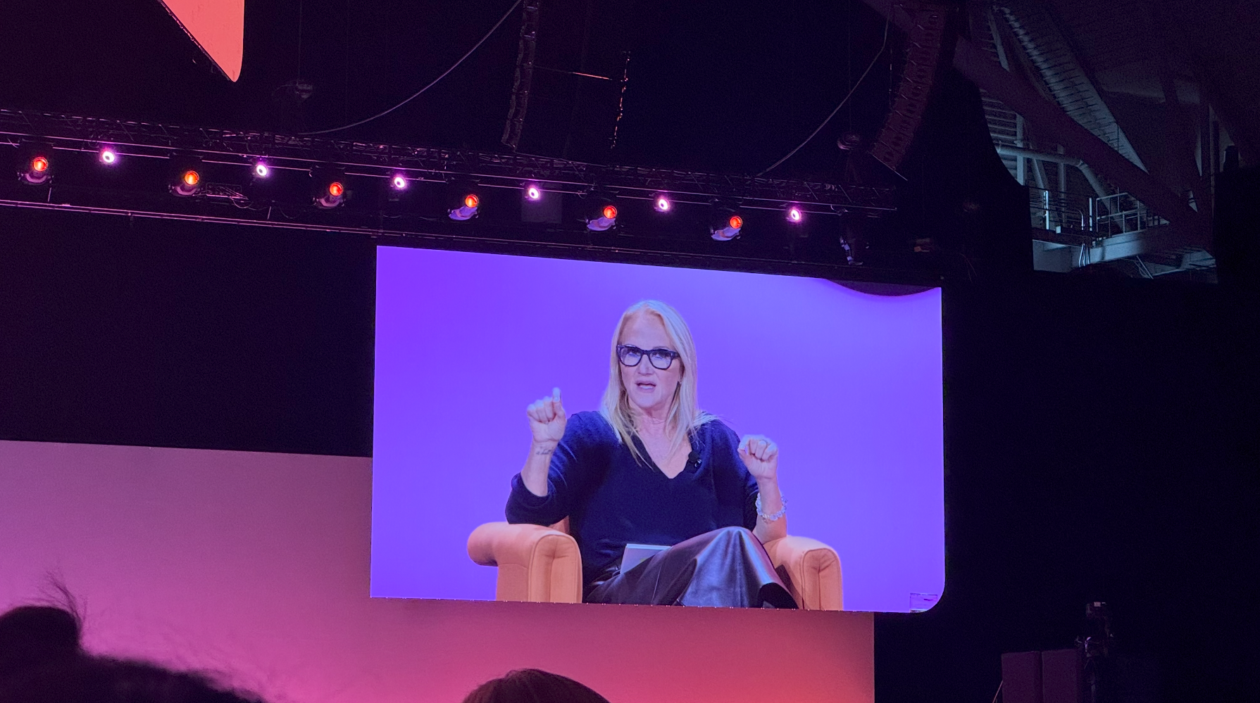 A large screen projection at the conference showing a woman seated in an orange armchair on stage
