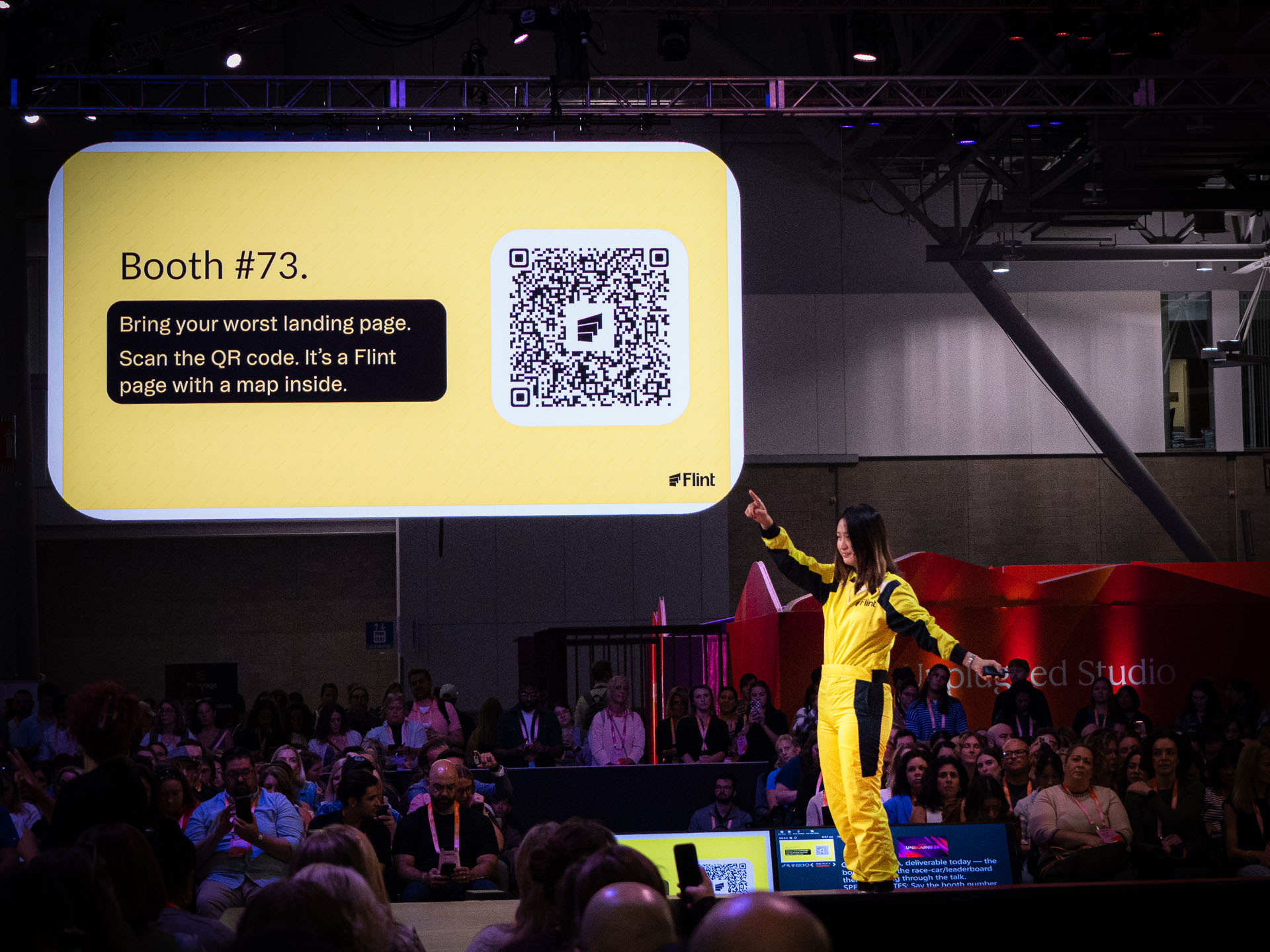 A crowded conference or event stage with a presenter in a yellow outfit pointing toward a large projected slide.