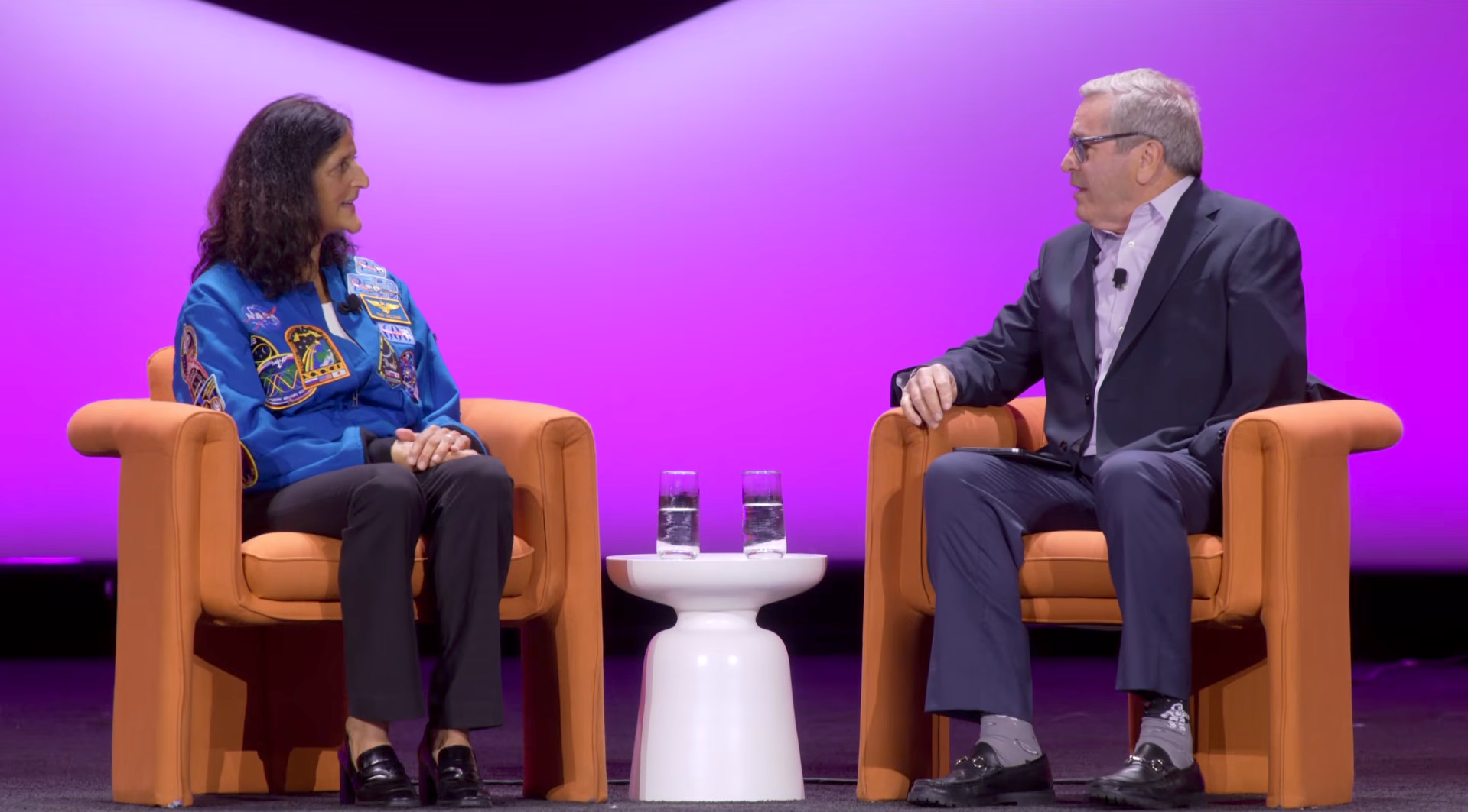 Two people seated on stage in armchairs facing each other and conversing, the person on the left wearing a blue jacket