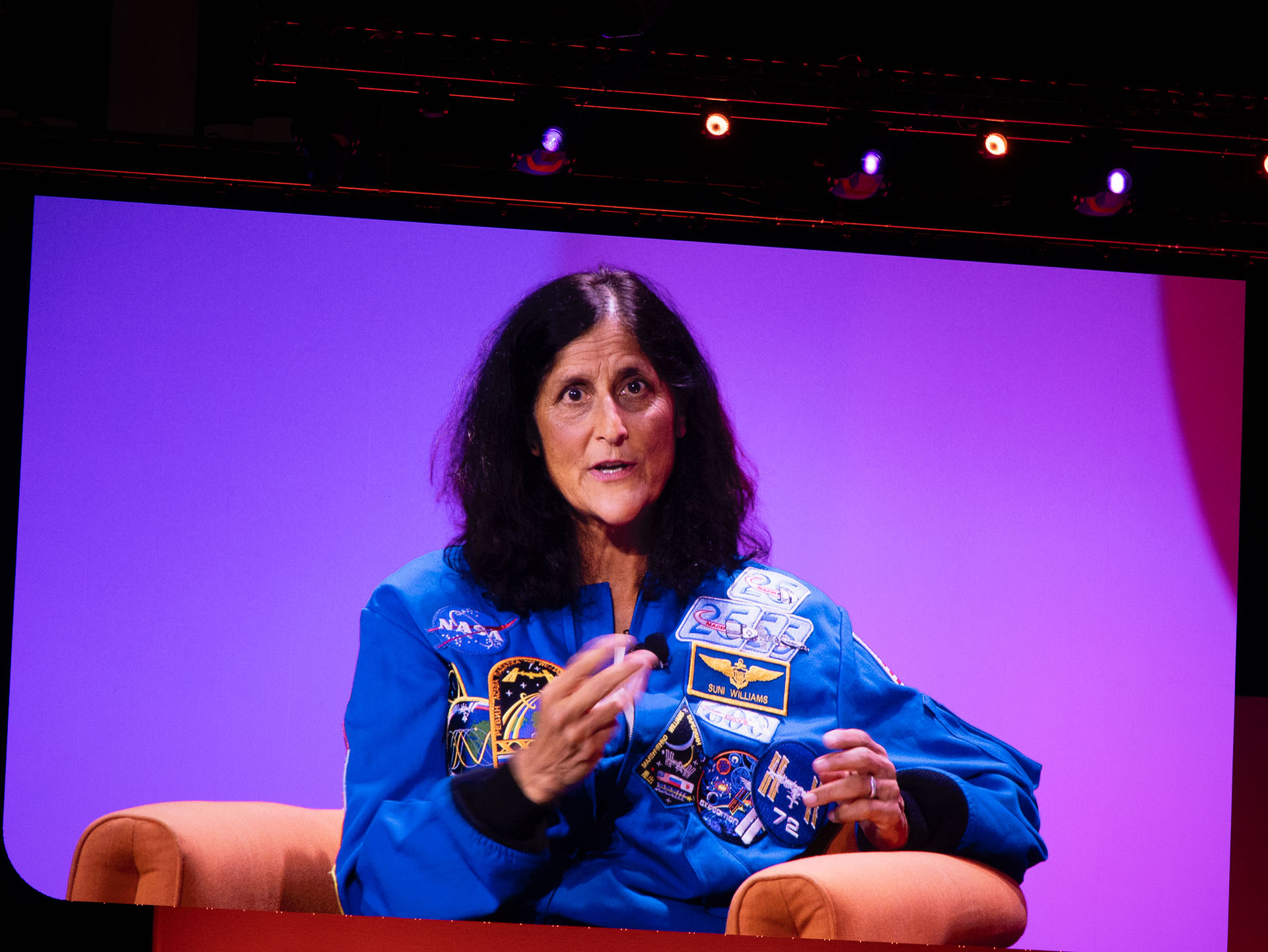 A woman seated on an orange armchair against a purple background, wearing a blue jacket covered with various mission patches