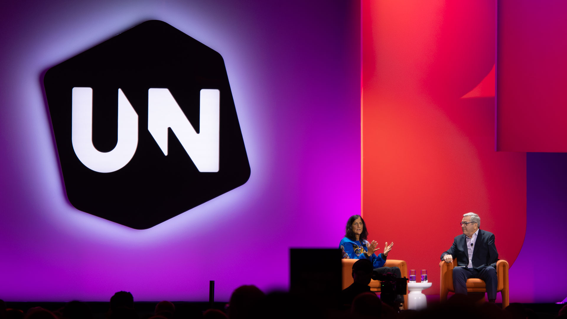 A stage presentation or panel discussion in a large venue with a purple and red gradient backdrop