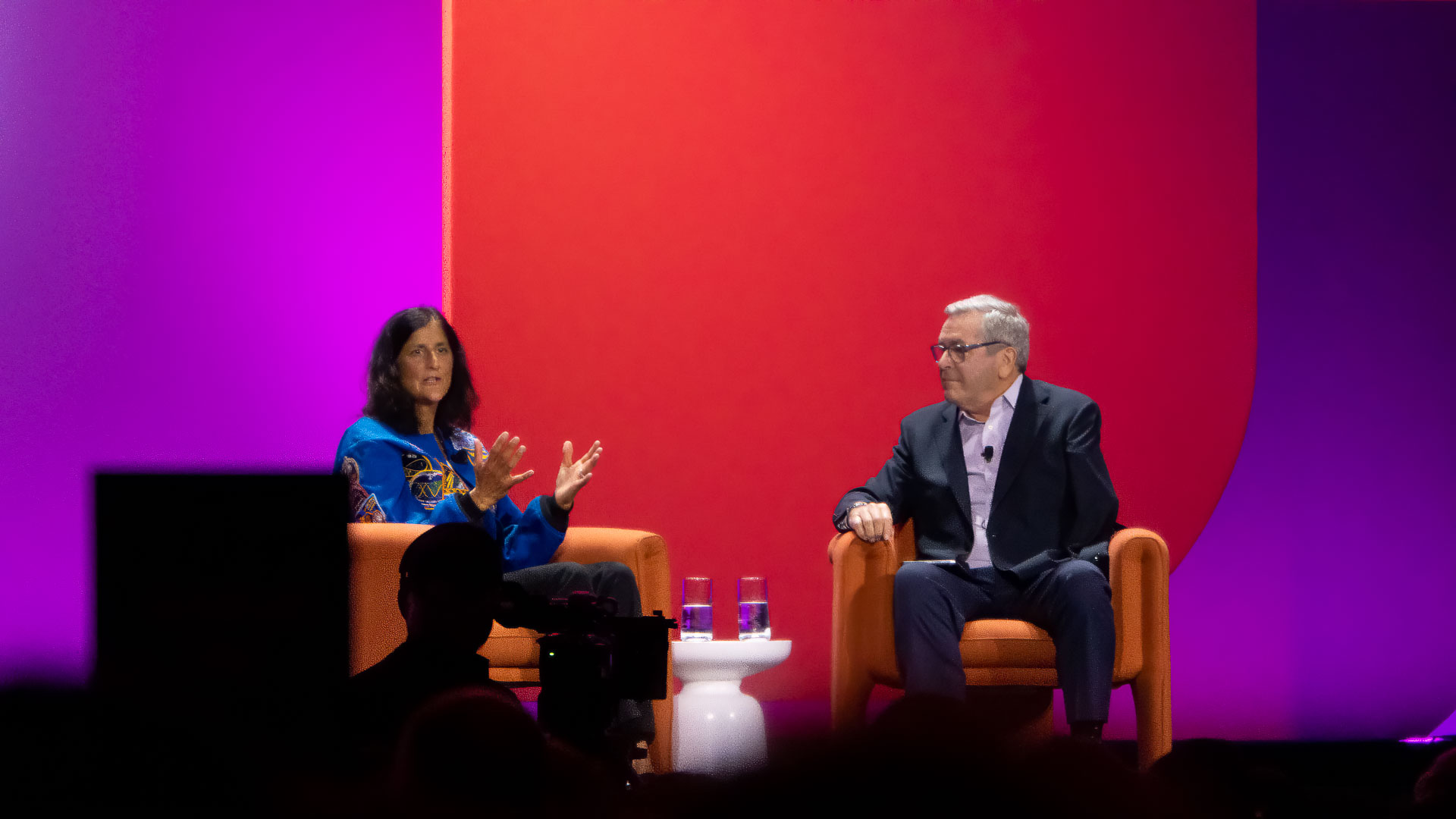 Two people seated on stage in armchairs against a vibrant purple and red backdrop, the person on the left gesturing while speaking