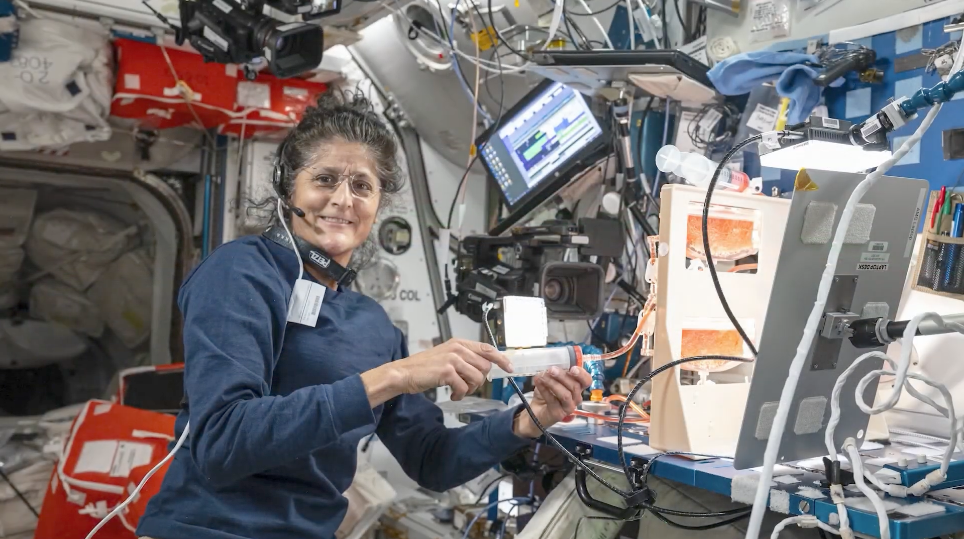 An astronaut inside a spacecraft or space station module, seated and smiling while working with equipment and cables