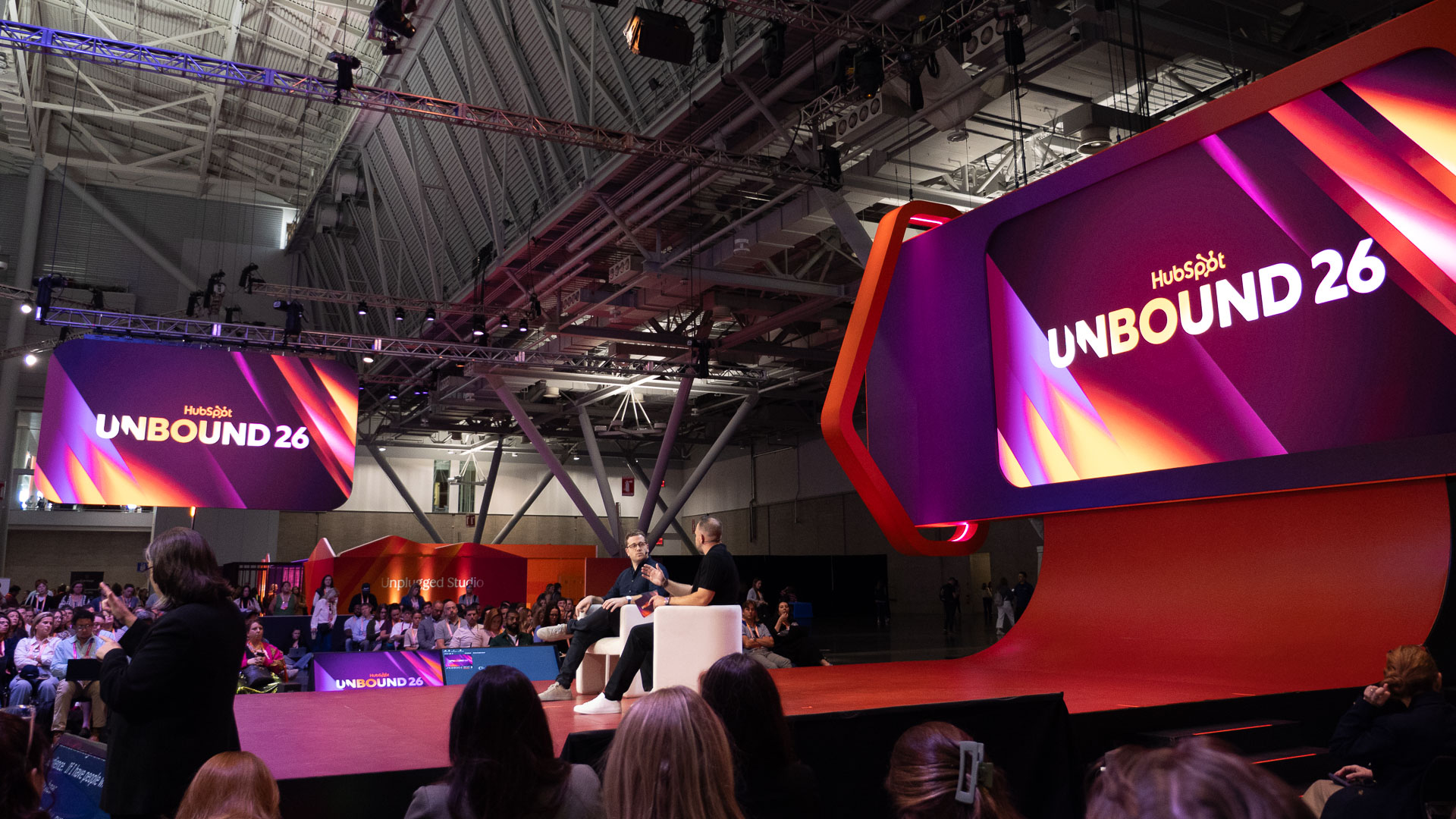 A large indoor conference stage with bright, colourful signage reading ‘HubSpot UNBOUND 26’ and two people on stage.