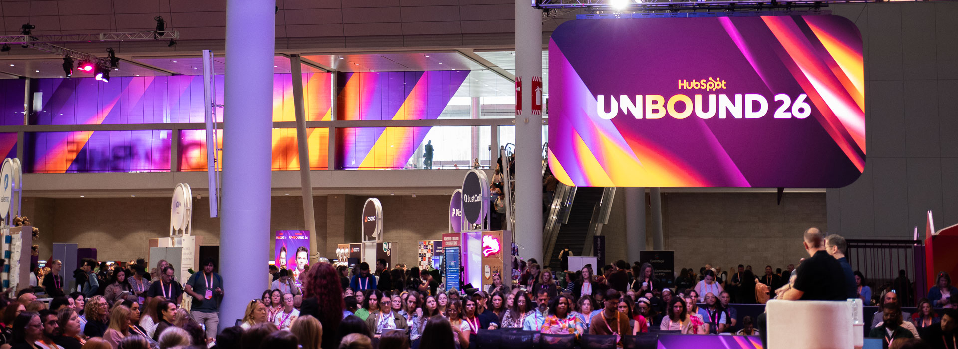 Panoramic establishing shot of a large indoor conference space with a stage at the right, where two speakers sit facing a seated audience.