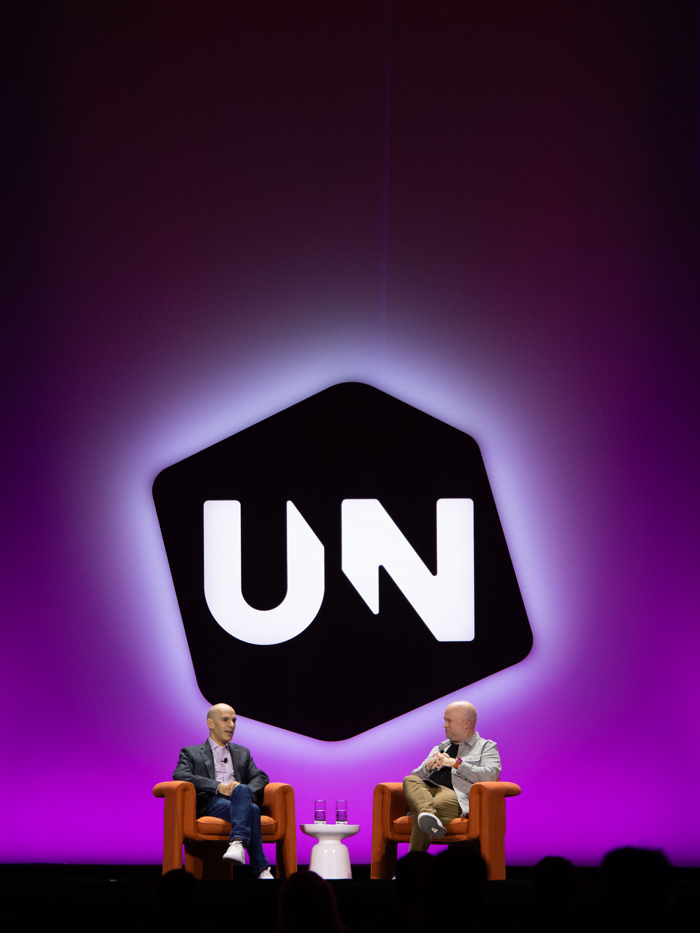 Two speakers seated in orange armchairs on stage in conversation in front of a large screen