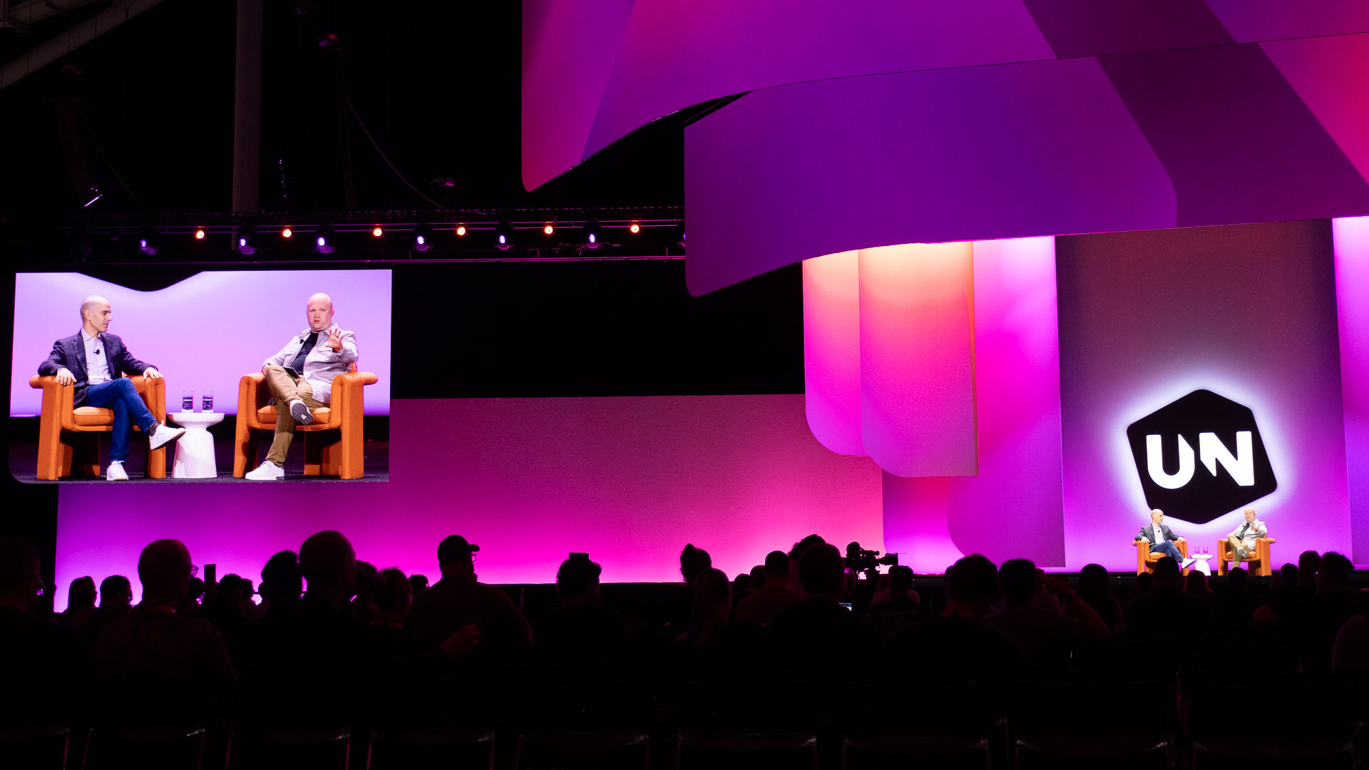 Stage panel discussion with two speakers seated in armchairs and their conversation projected on the screen behind them