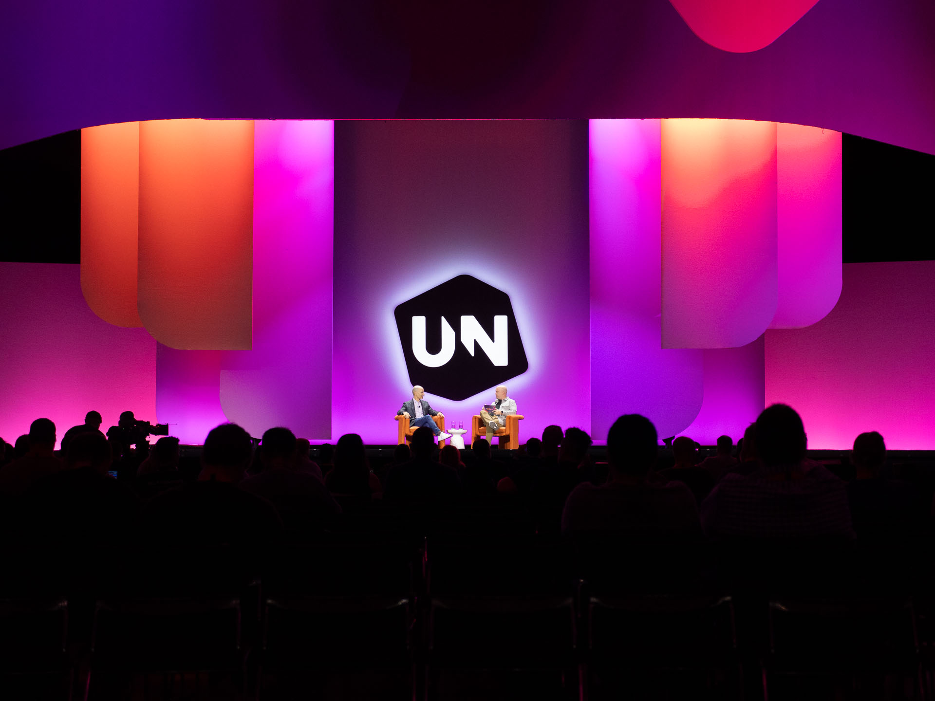 Two speakers seated in chairs at the centre of a conference stage under pink, purple and orange lighting