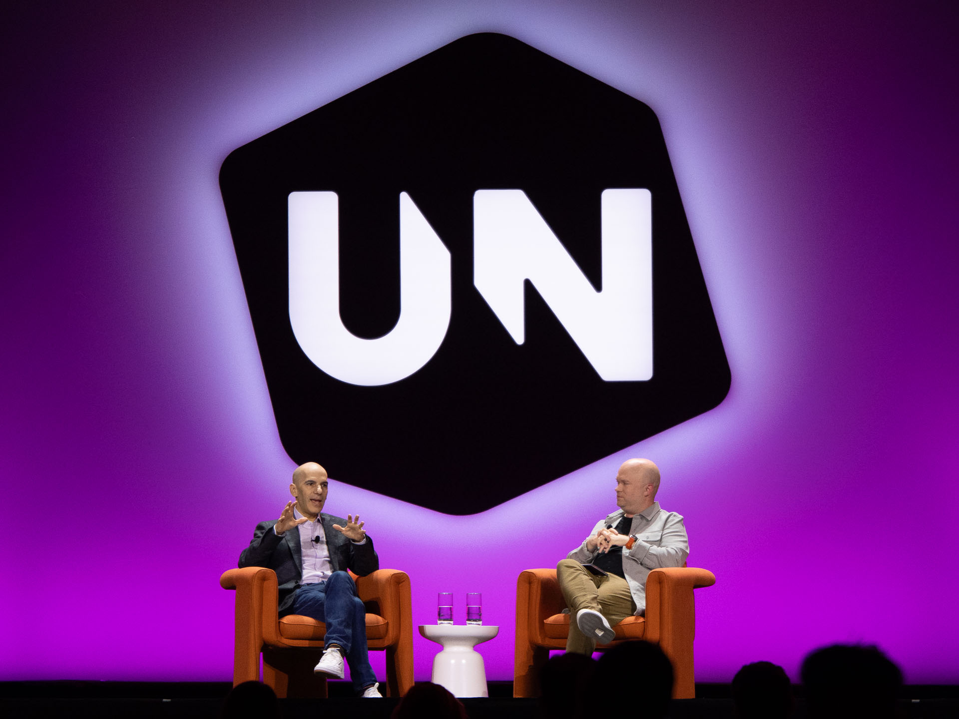 Two speakers on stage in orange armchairs against a vivid purple and pink background with a large logo behind them