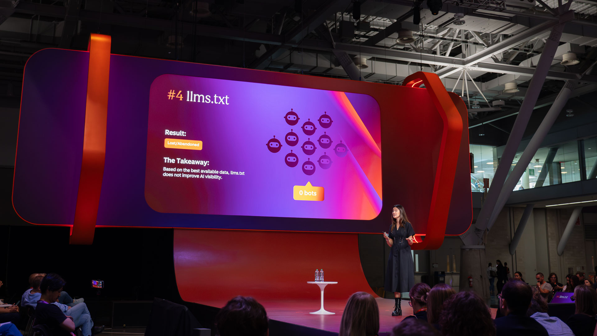 Wide-angle view of the session stage in a large indoor venue, with a curved red and purple display screen behind the presenter