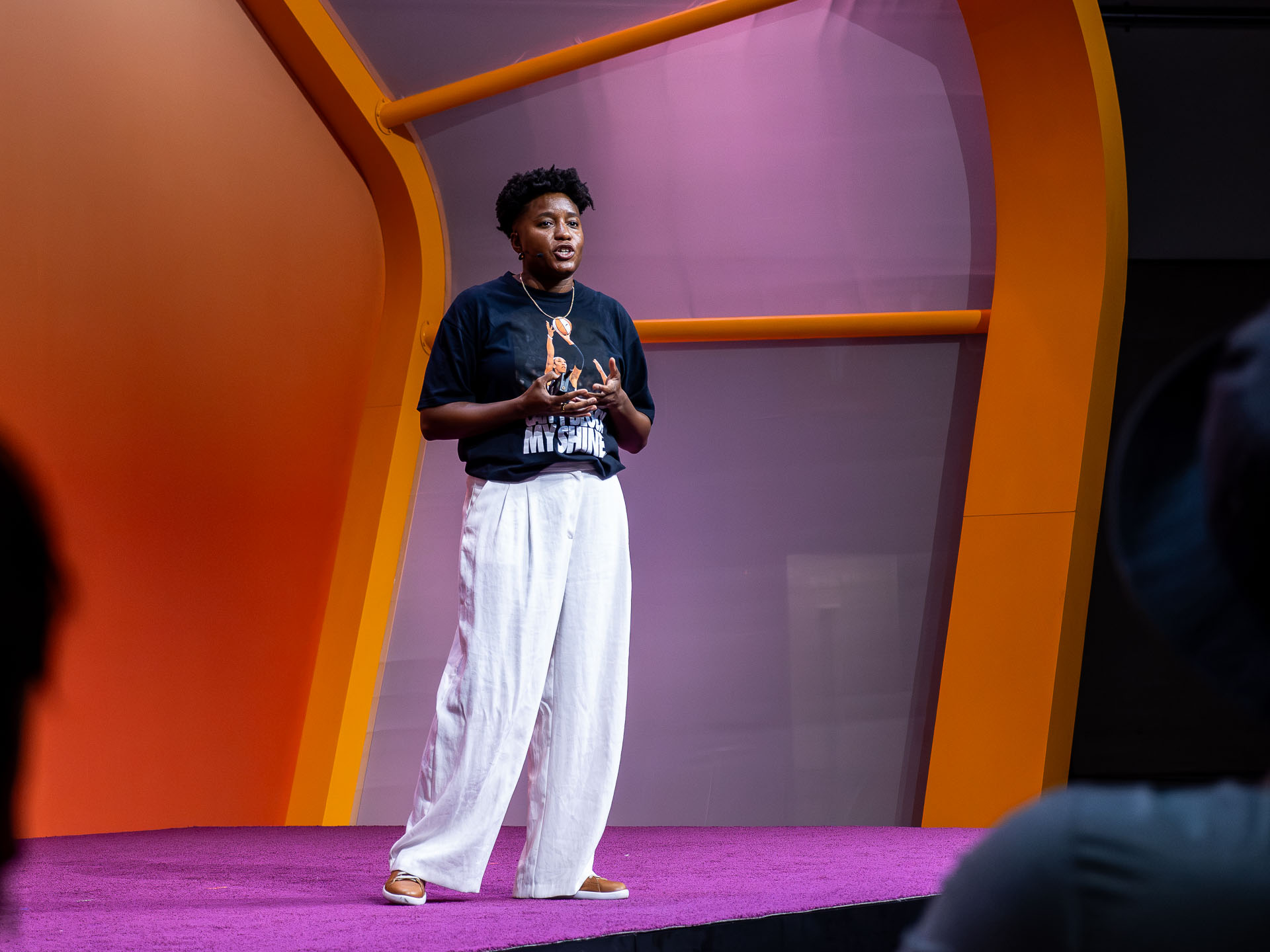 The speaker on a purple-carpeted stage, positioned slightly left of centre and wearing a black graphic t-shirt while presenting