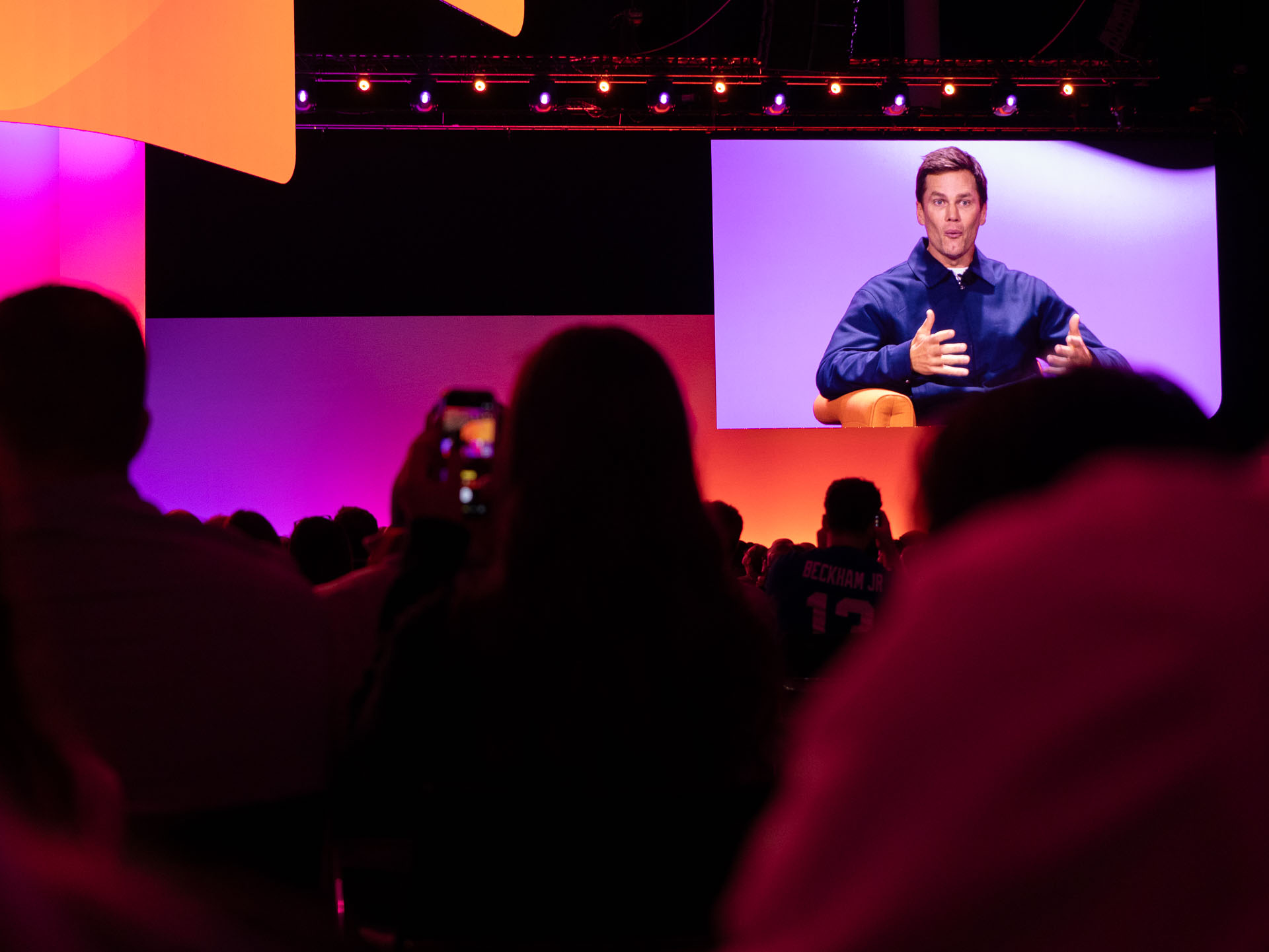 Indoor conference with an audience seated in the foreground, mostly silhouetted against coloured stage lighting