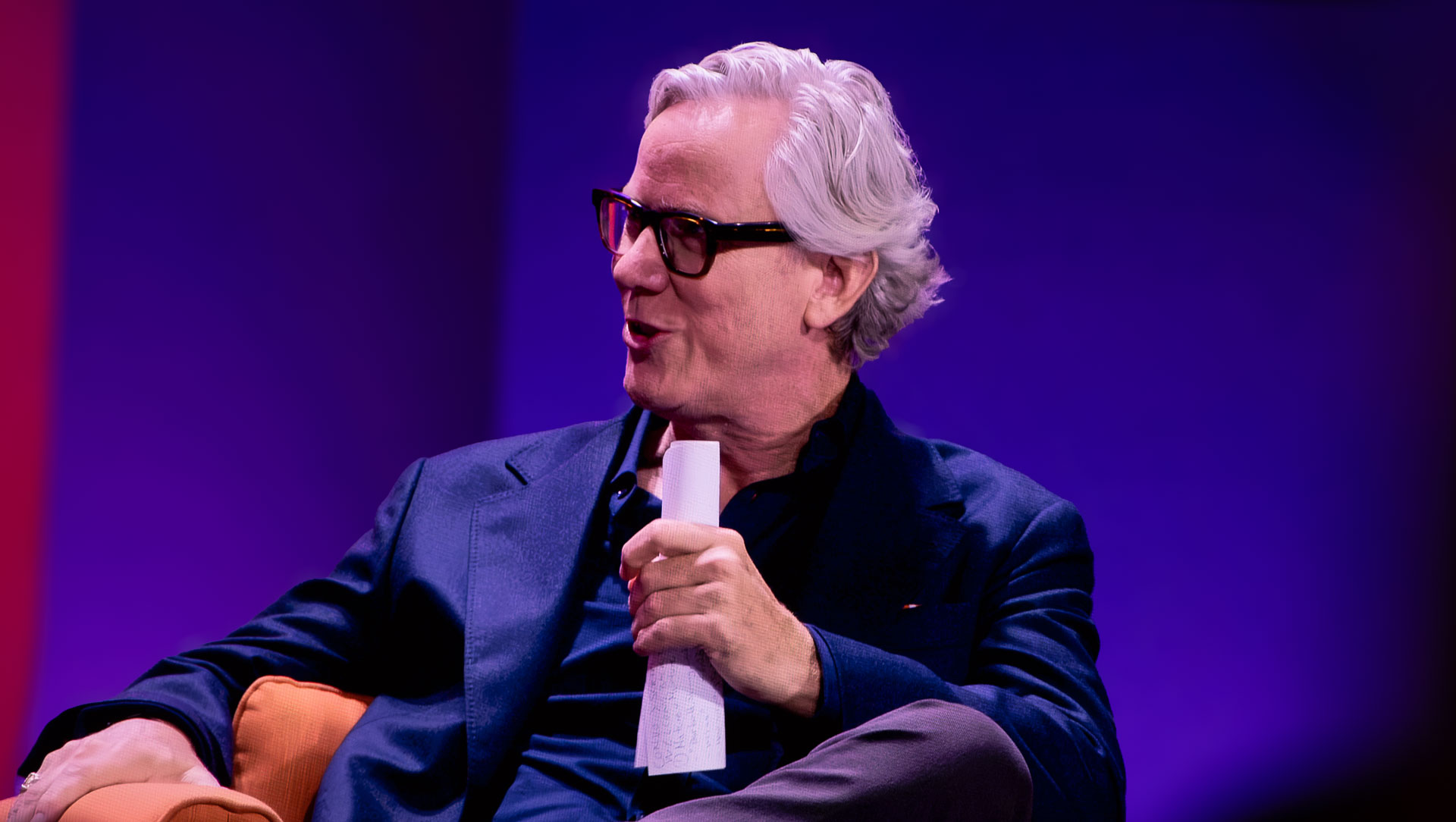 An older man with grey hair and black-rimmed glasses seated on stage against a purple-lit background