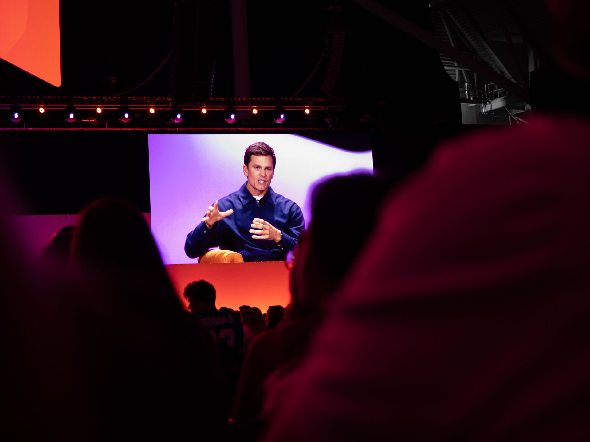 Large screen at an indoor event projecting a speaker wearing a dark shirt who is gesturing with his hands