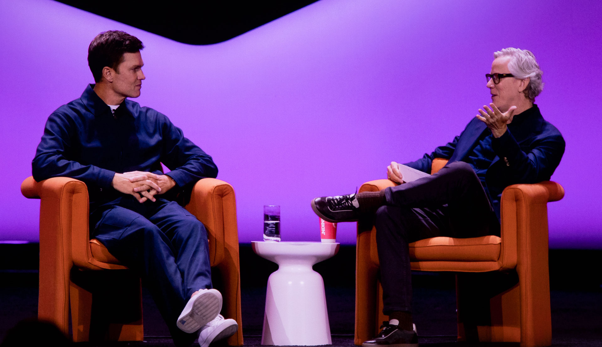 Two men sit on stage in conversation against a purple-lit backdrop, each in an orange armchair with a small white round table