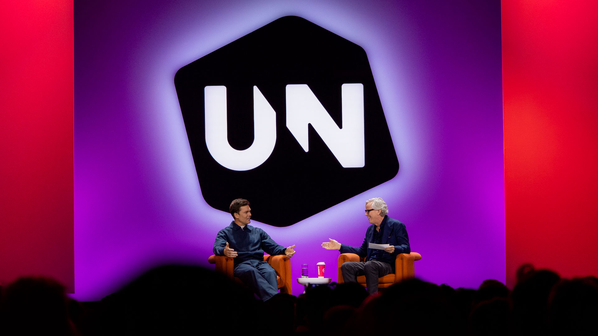 Two men seated on stage in armchairs engaged in a conversation or interview, with a large stylized logo behind them