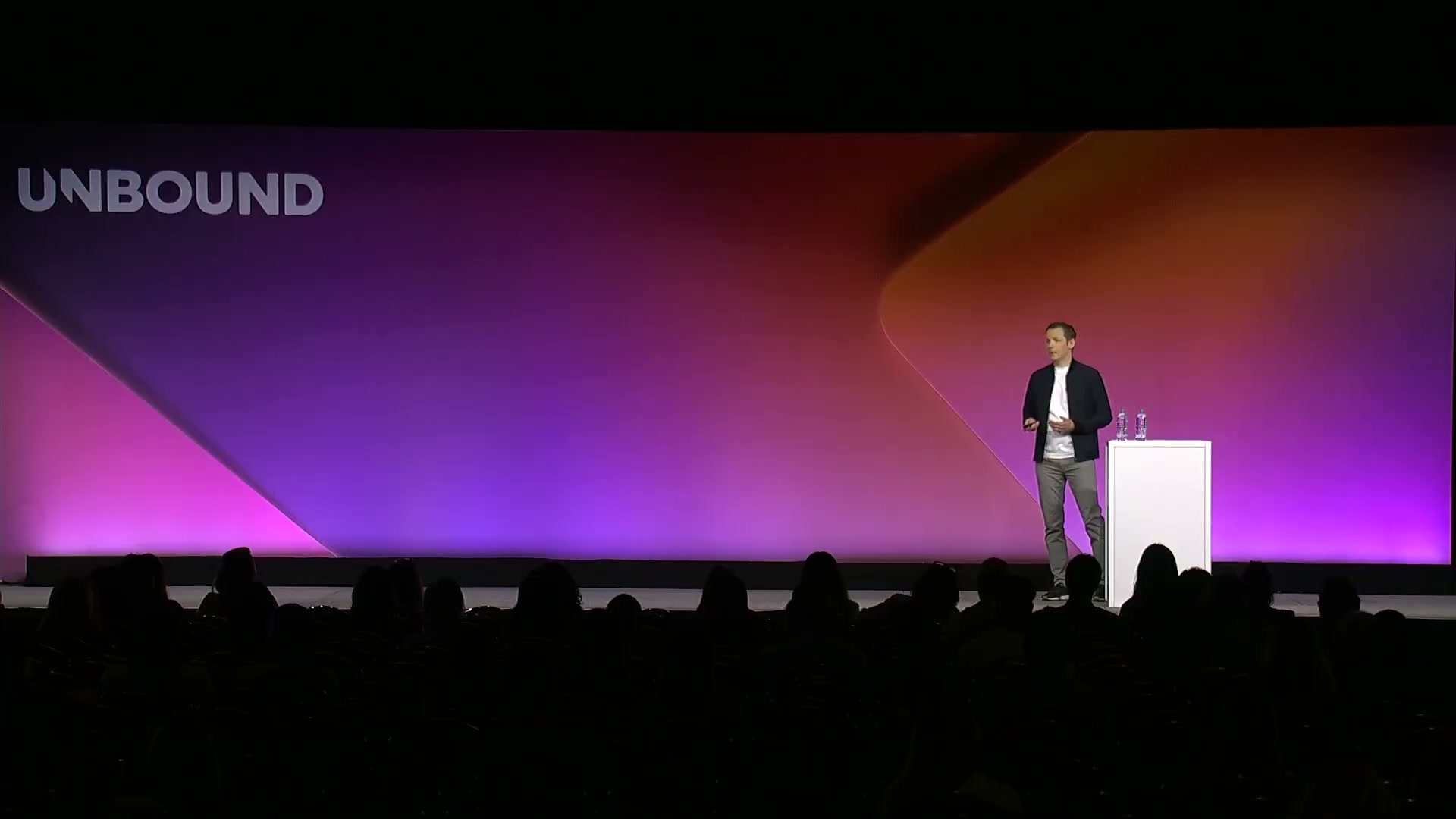 Wide-angle view of a dark auditorium with a single speaker standing to the right of the stage behind a small white podium