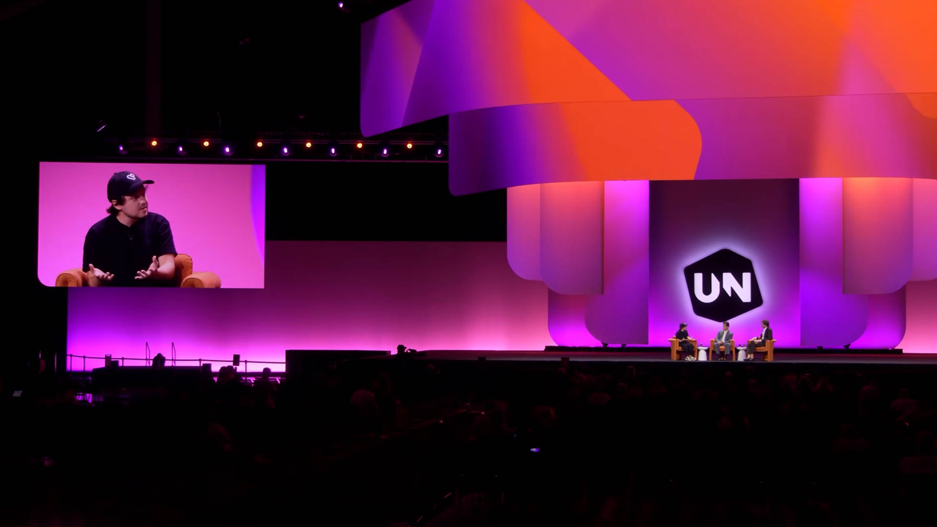 Wide view of the UNBOUND auditorium, with three people seated in chairs on the stage under purple and orange lighting
