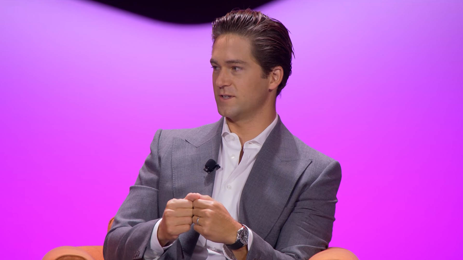 A man in a grey suit and white shirt seated on an orange chair against a pink and purple gradient background, turned slightly to one side