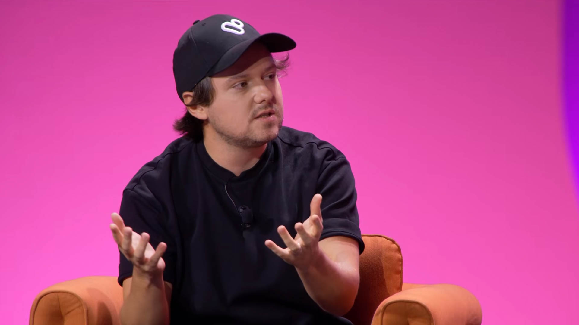 A man in a black jacket sits on an orange armchair against a pink background, gesturing with both hands as he explains a point