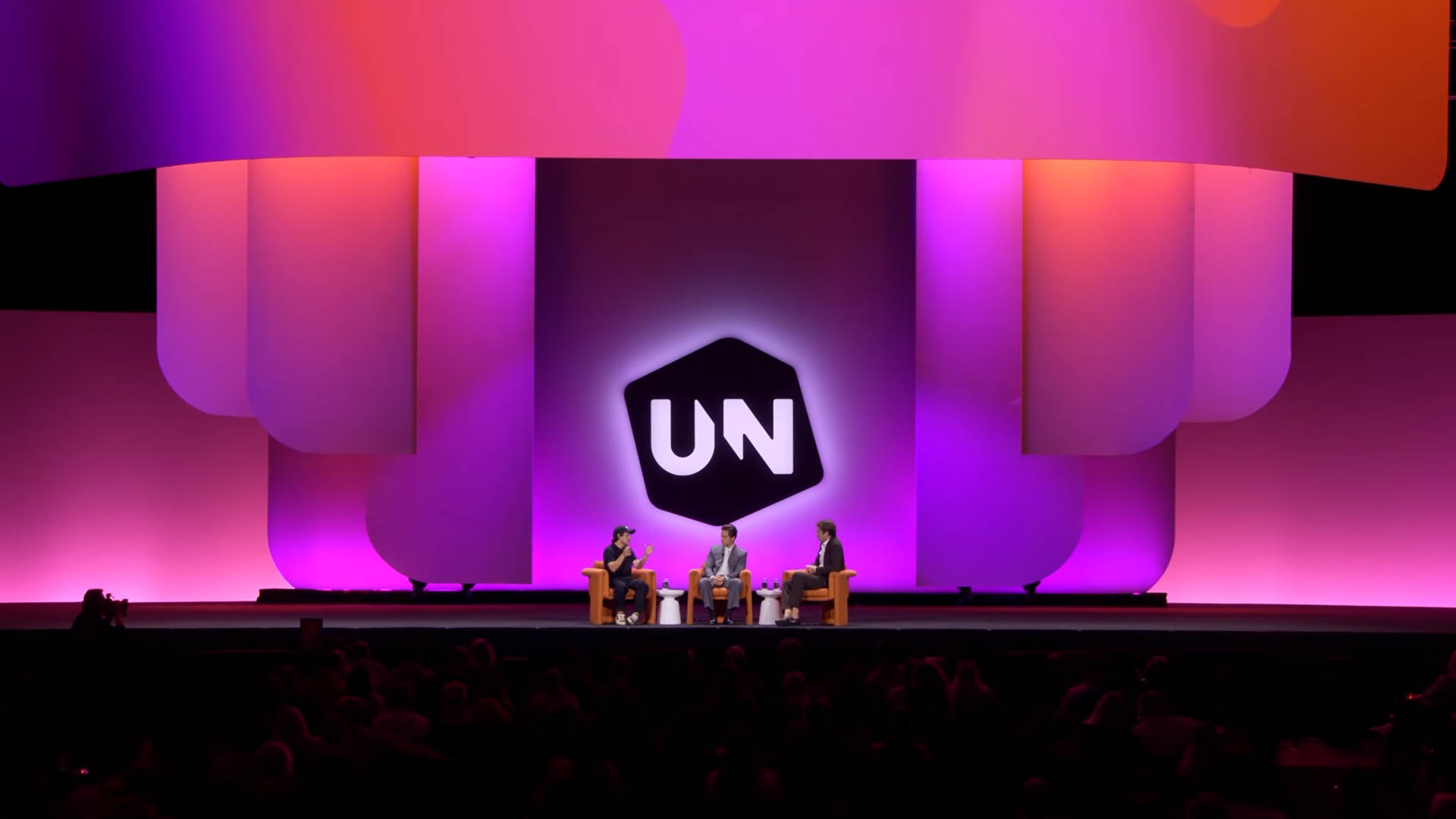 A stage set for a panel discussion with three people seated in armchairs under magenta and purple lighting, with a large stylised backdrop
