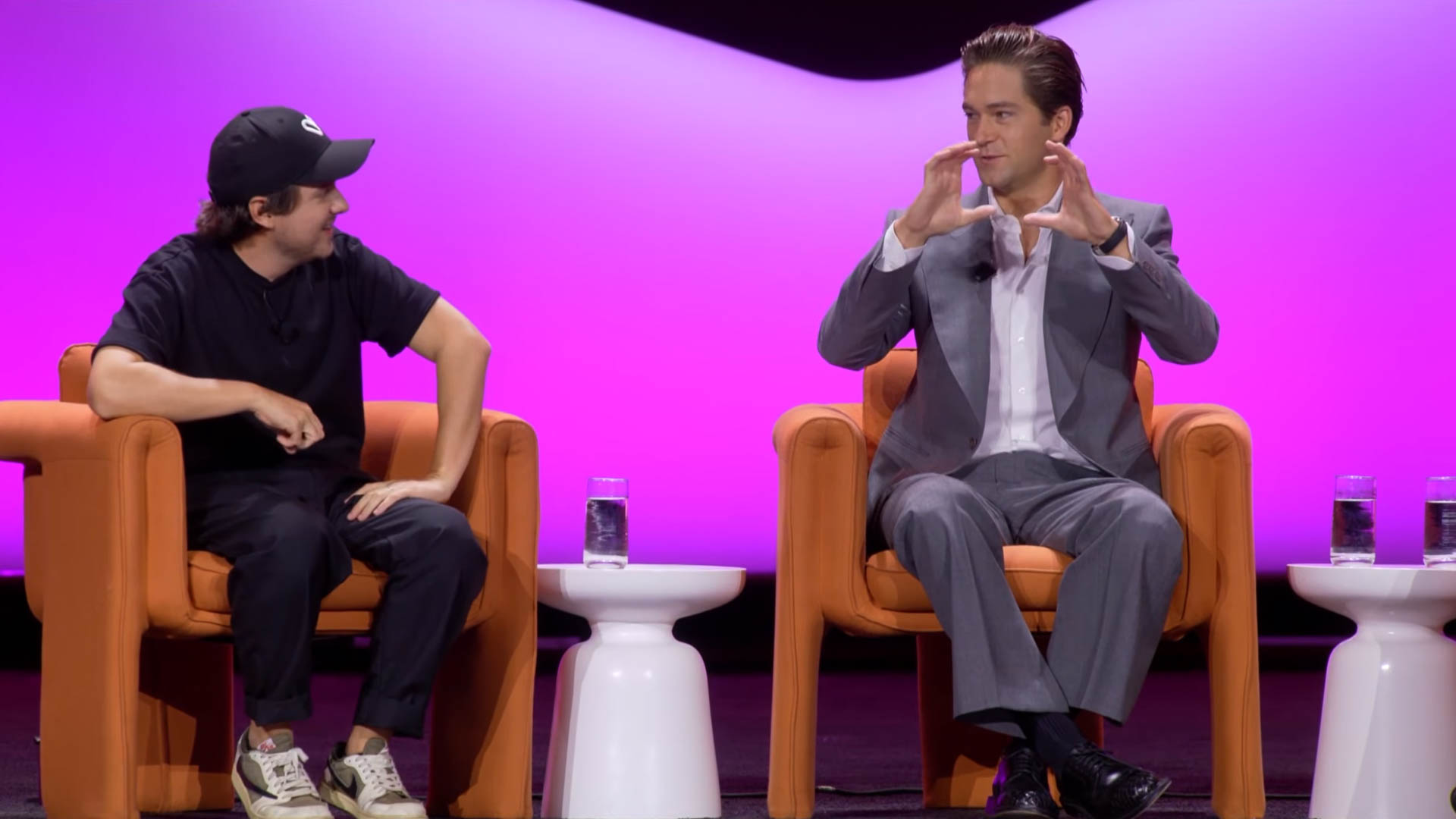 Two men seated on stage in orange armchairs against a purple and pink gradient backdrop, the man on the left wearing a black jacket