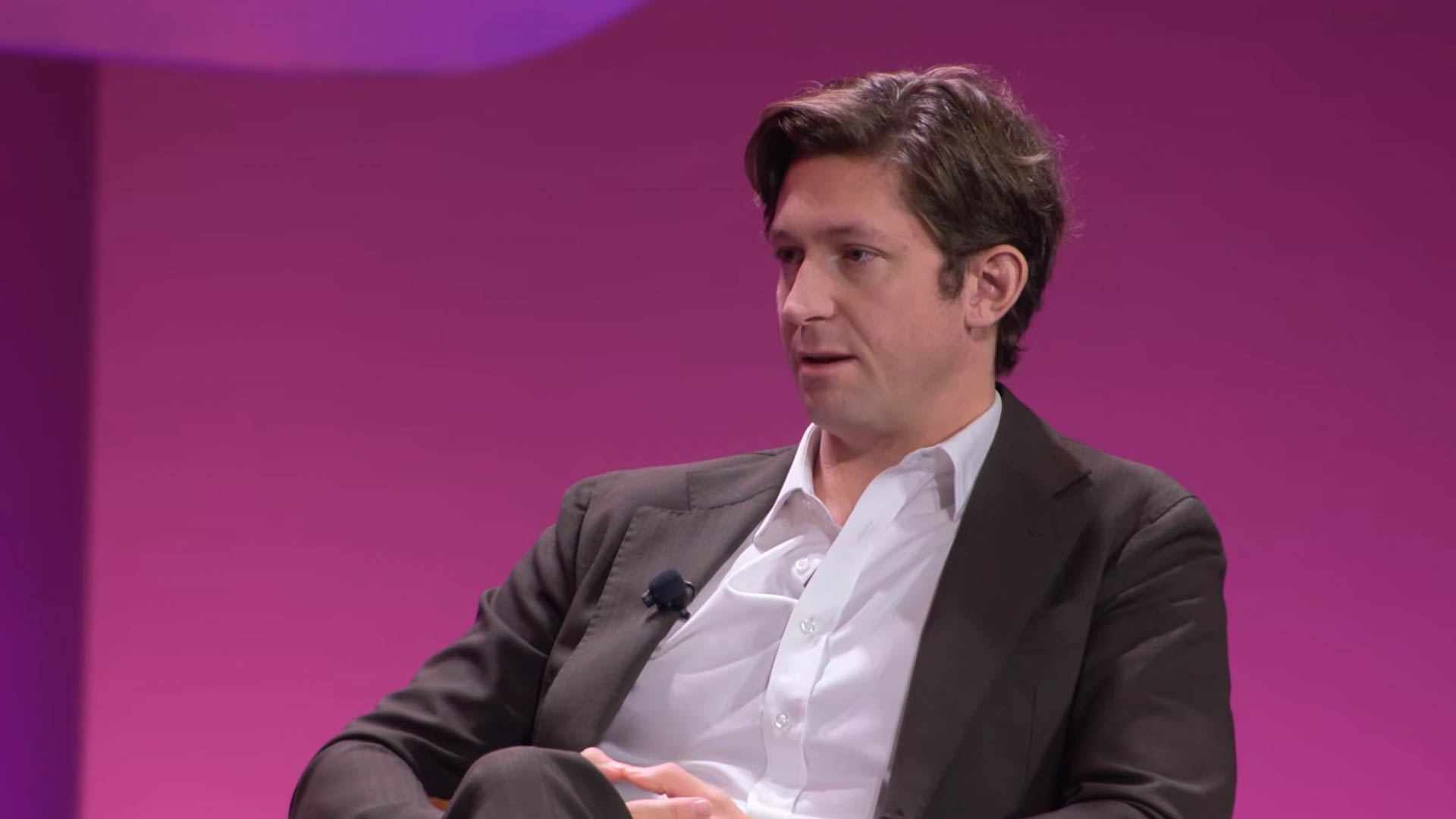 A man with short brown hair in a dark suit jacket over a white dress shirt, seated against a pink background